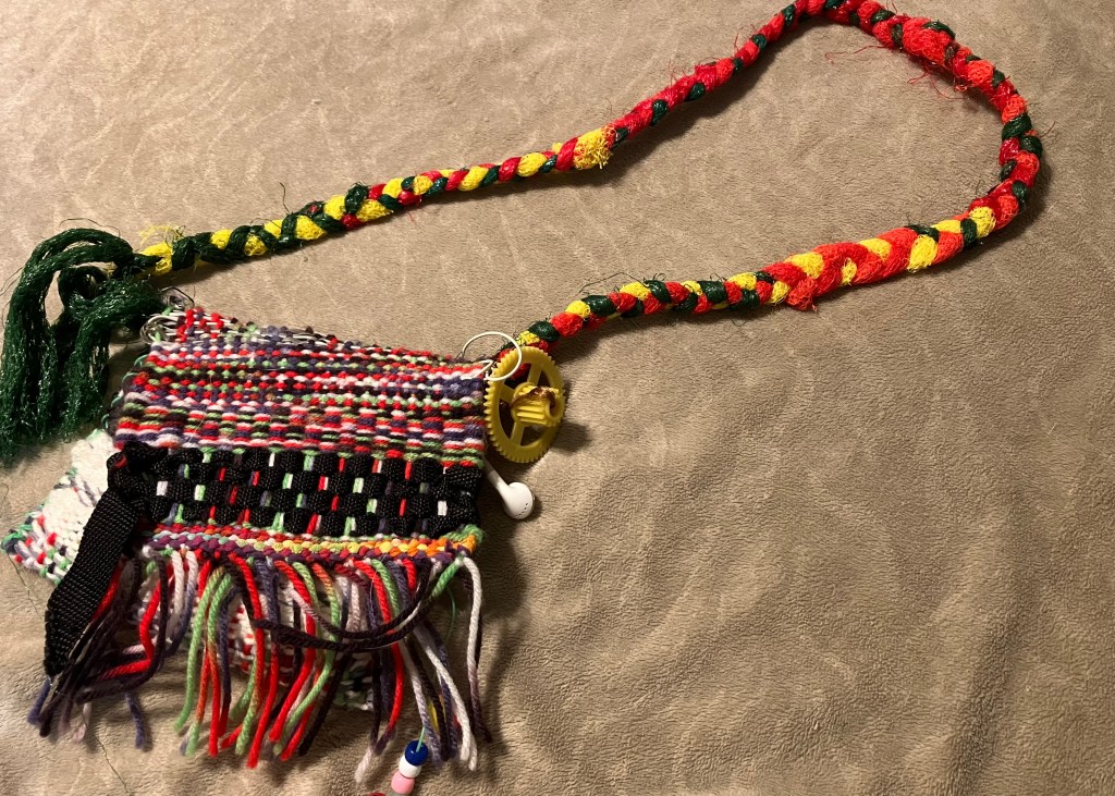 A small, multicolored woven bag made by fiber artist, Sarah Batchelder. The bag is woven from an assortment of fibers, including a black nylon strap and an old pair of earbuds. A plastic gear is attached for decoration.