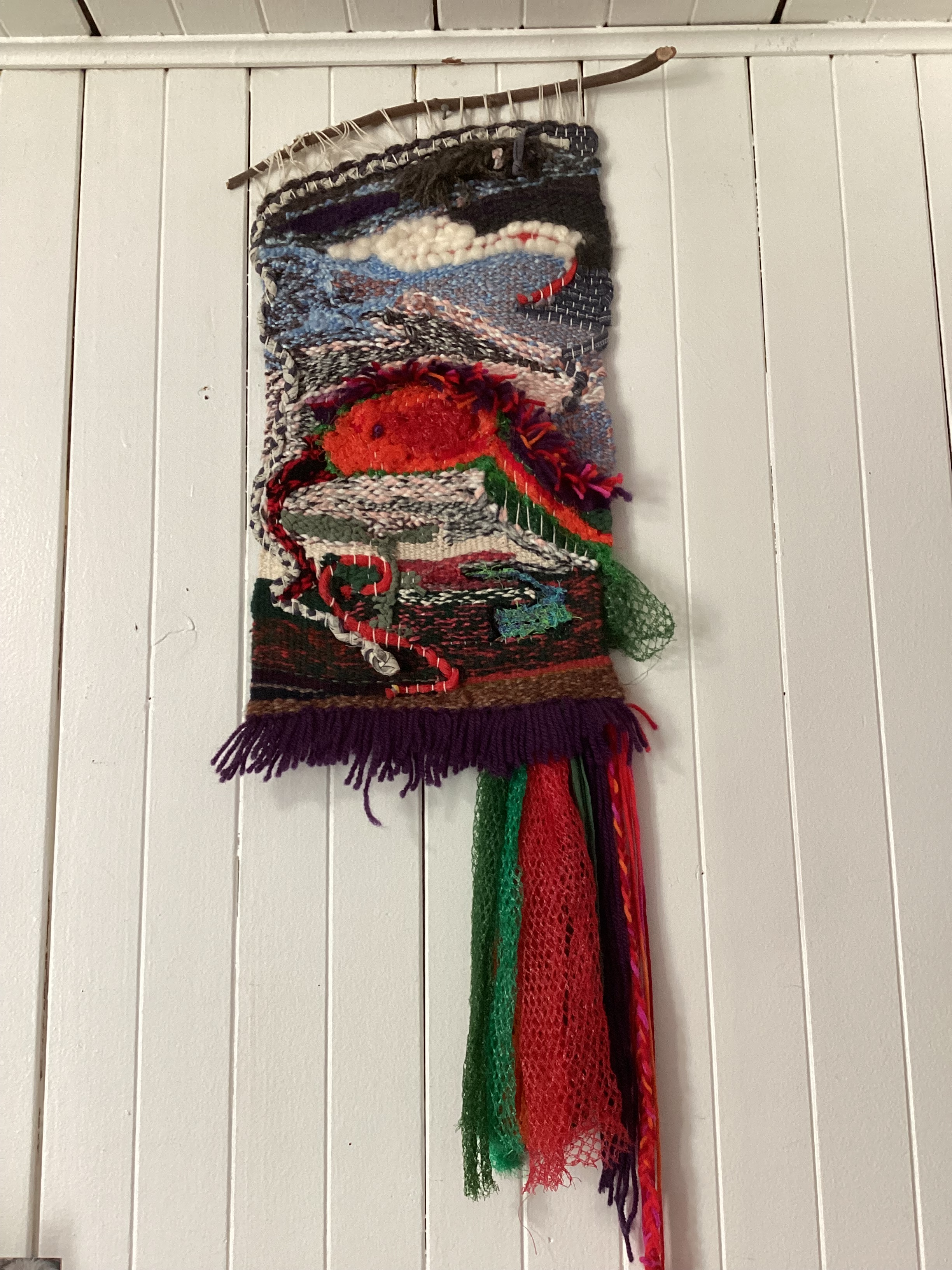 A handwoven tapestry by fiber artist, Sarah Batchelder, depicting a large snake head (with fringe) and other smaller snakes in the sky and earth.