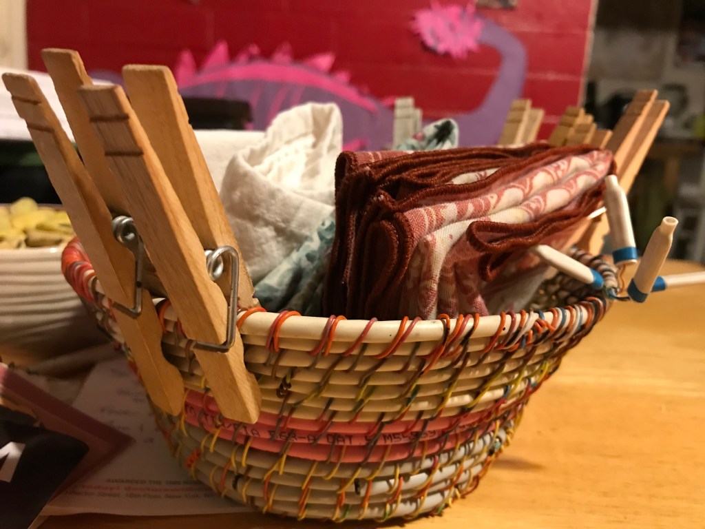 A household basket made by Sarah Batchelder from repurposed telephone and cable wires. It holds cloth napkins and stores clothespins around the edges.