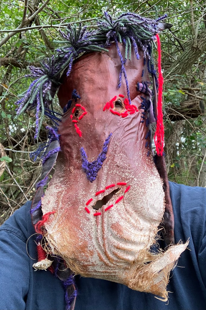 Fiber artist, Sarah Batchelder, posing in a hand stitched mask made from palm fiber, yarn, and found beans.