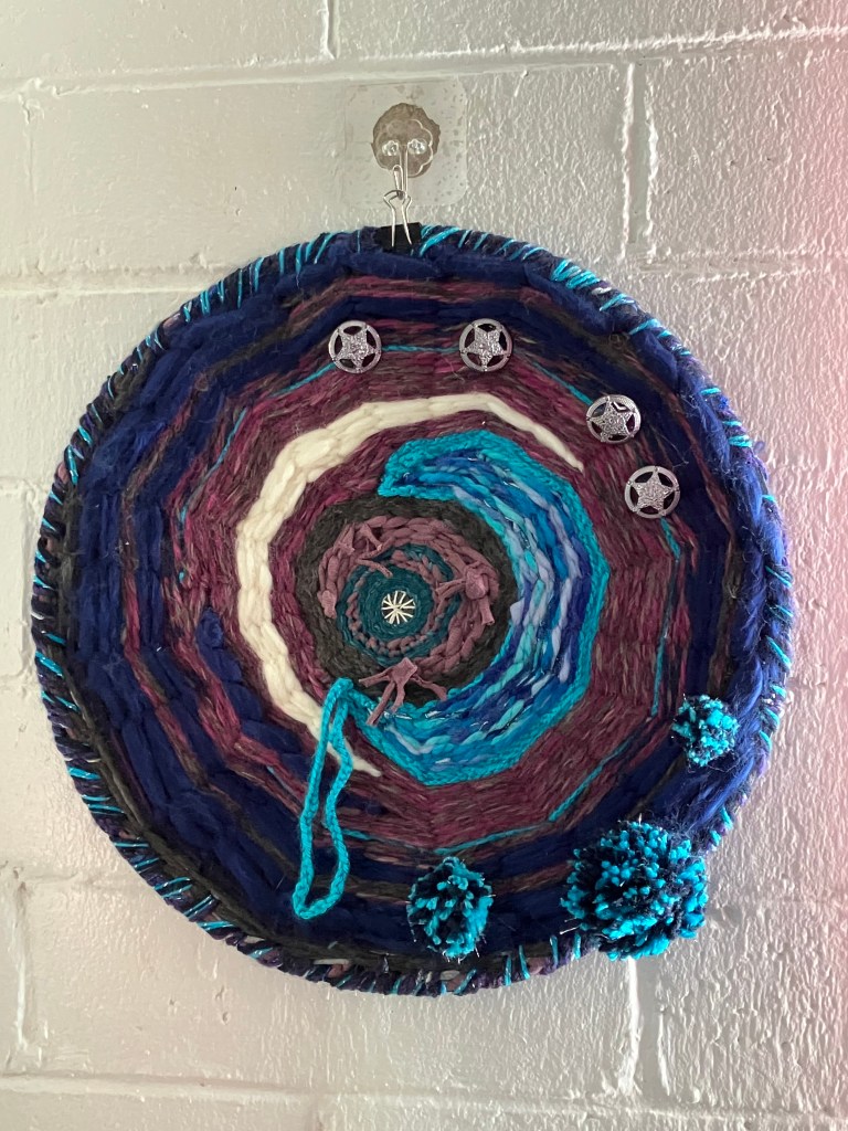 A circular weaving by Sarah Batchelder depicting two moon shapes with button stars and celestial pom poms.