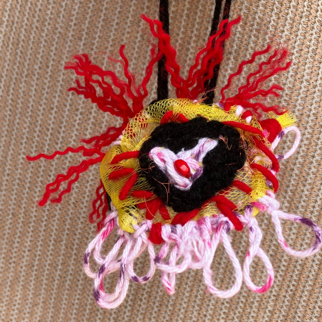 A hand-stitched mini hanging of an eyeball. A red bead is surrounded by a heart on a black and yellow background.